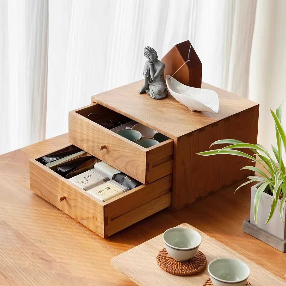 A light natural wood multi-drawer organizer with two open drawers, styled on a wooden table with tea cups, a small statue, and a plant in a home interior setting.
