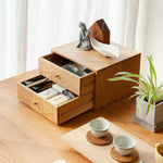 A light natural wood desk organizer with two open drawers and a top shelf, styled on a wooden table with tea cups, a small plant, and decorative objects in a bright, clean interior setting.