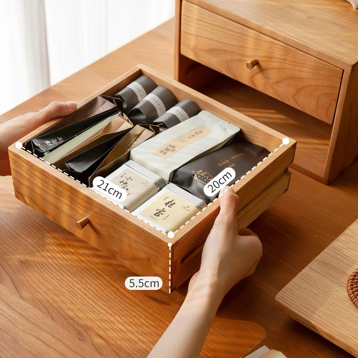 A wooden drawer organizer being held open by two hands, showing compartments filled with packaged tea or coffee bags. The organizer is shown on a wooden table, with a matching wooden drawer unit in the background.