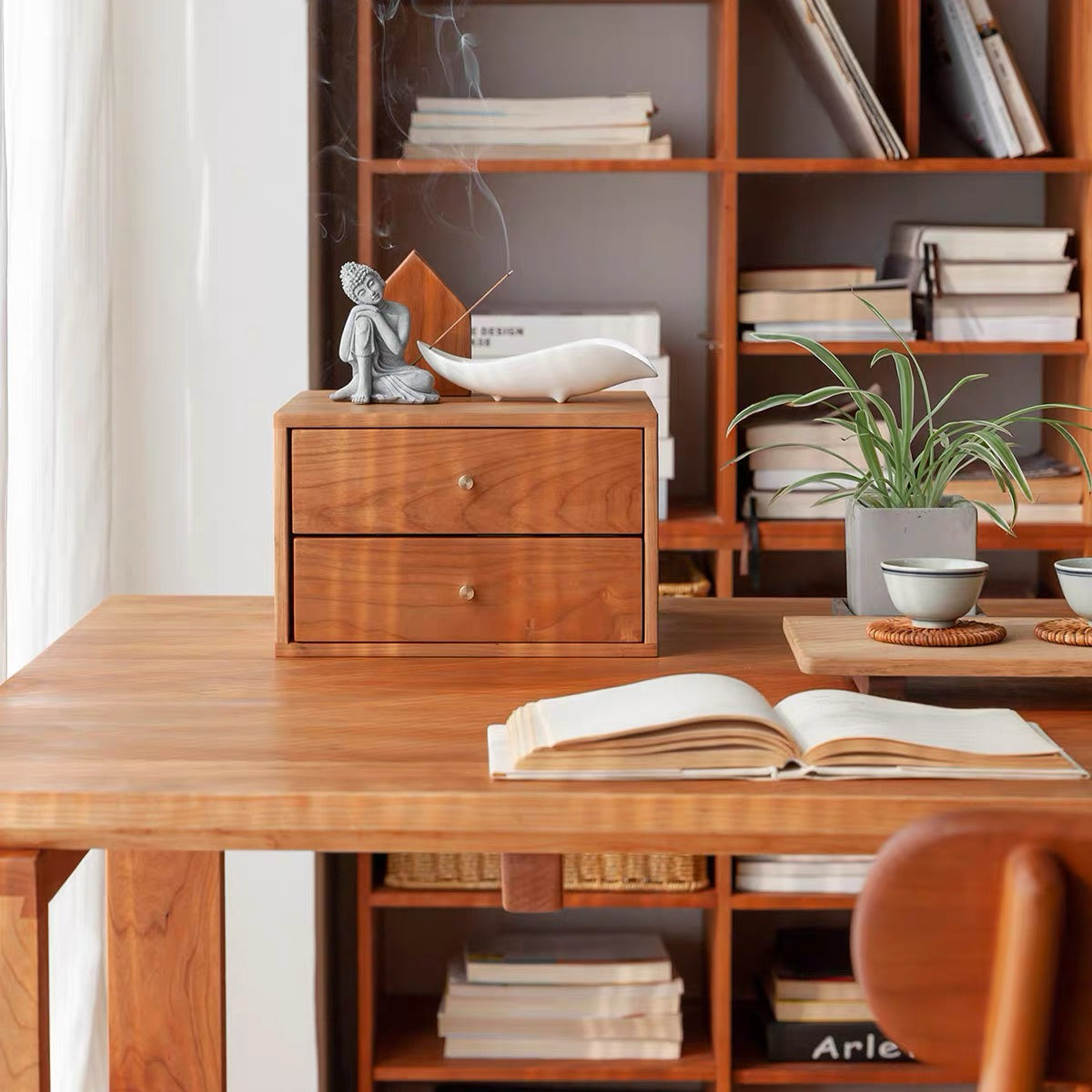 A warm-toned wooden two-drawer desk organizer box sits on a wooden table in a styled home office or study setting, with books, a small plant, and an open book nearby.
