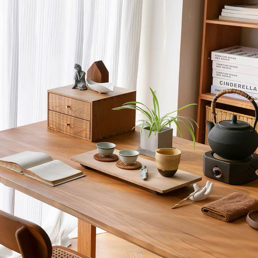 A styled wooden tea set and table accessories arranged on a warm wood desk in a bright room. Visible items include a black teapot on a small electric burner, ceramic cups and bowls on a tray, a potted plant, books on a shelf, and a small figurine.
