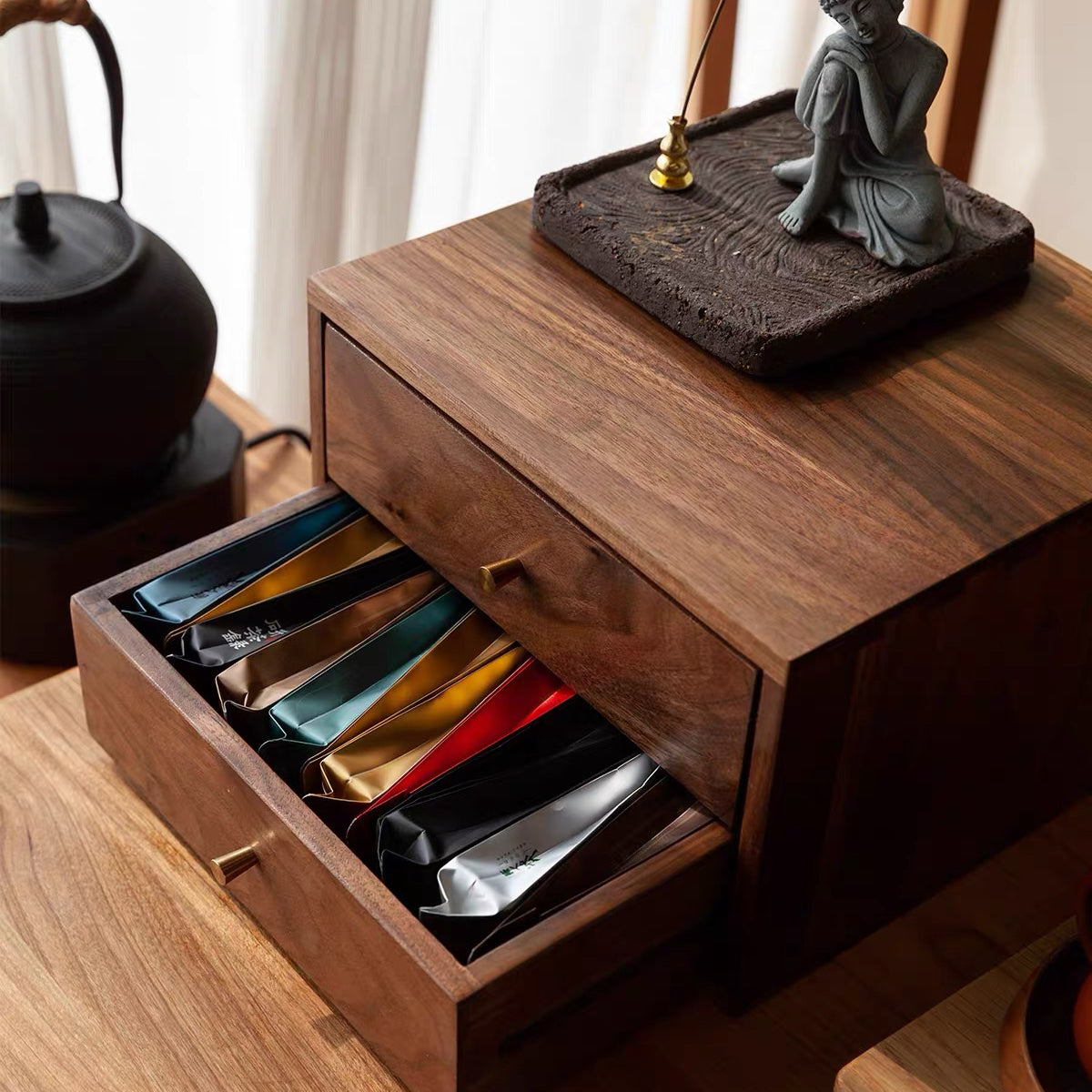 A dark wood drawer organizer with one open drawer containing colorful tea bags, styled on a wooden table next to a black teapot with a decorative statue on top.