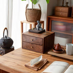 A dark brown wooden two-drawer storage cabinet with brass knobs sits on a wooden table in a styled home interior. A small black figurine and thin rod rest on top of the cabinet.