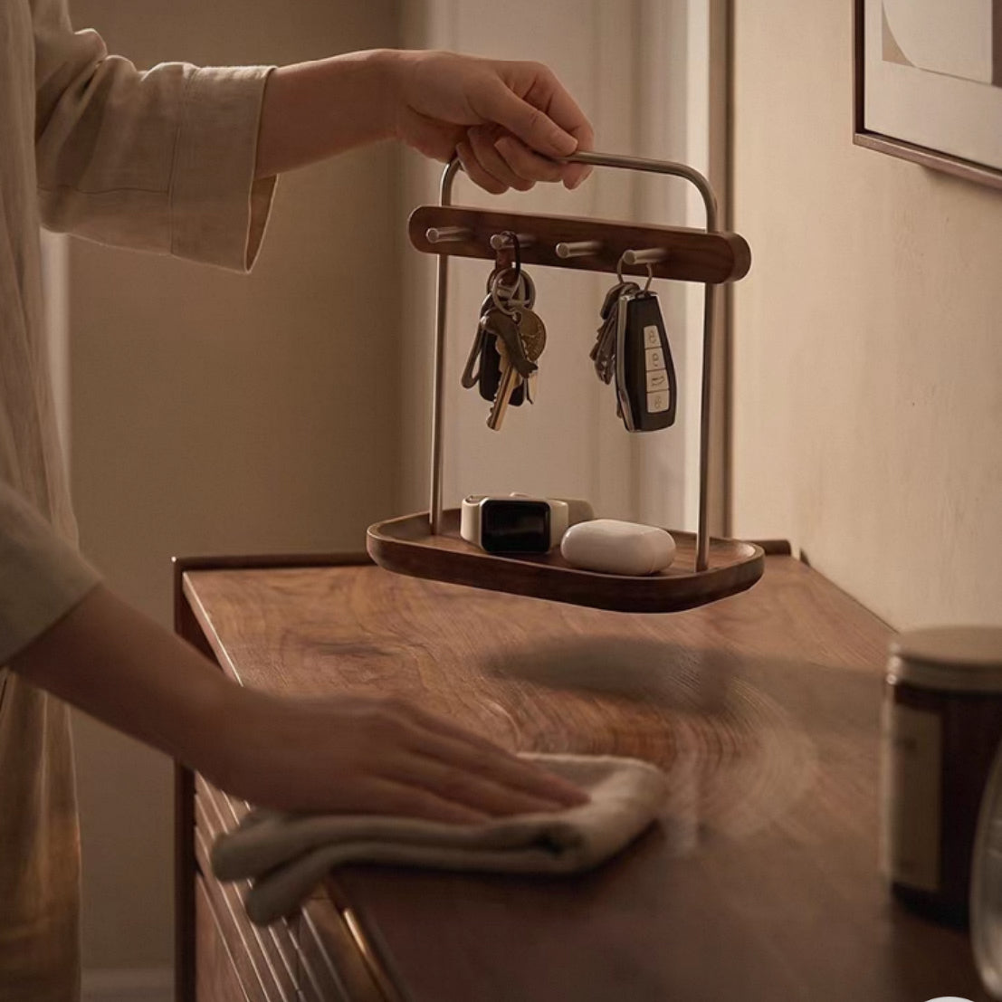 A wooden and metal key holder tray with a curved top handle is being held by a person and placed on a wooden cabinet. Keys hang from the top hooks, and a small tray dish below holds a watch and a white oval item.