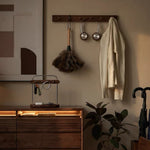 A dark wood wall-mounted coat rack in a styled interior, holding a cream knit sweater, silver over-ear headphones, and a dark feather duster. A matching wooden drawer unit with warm under-light sits below, alongside a plant and a folded umbrella.