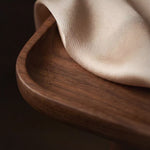 A close-up of a cream-colored textured fabric draped over the curved rim of a dark brown wooden tray against a dark background.