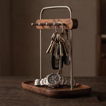 A wooden and metal key holder stand on a dark wooden tray, holding a set of keys and a wristwatch, photographed on a dark wooden table against a warm brown background.