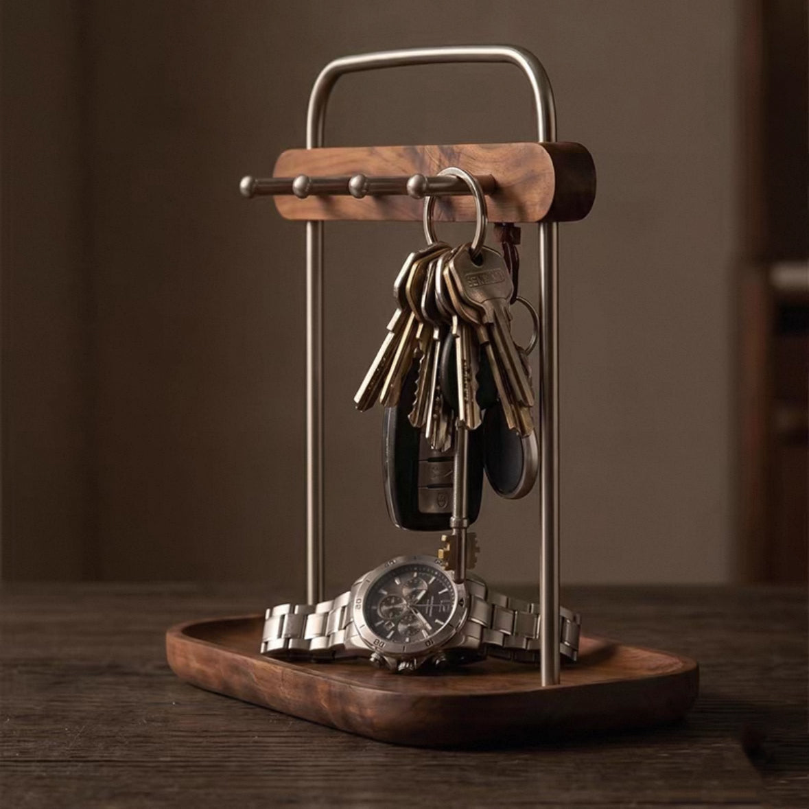 A wooden and metal key holder stand on a dark wooden tray, holding a set of keys and a wristwatch, photographed on a dark wooden table against a warm brown background.