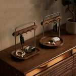 Two wooden key and accessory organizers with metal stands sit on a wooden dresser in a warm, styled room. The left organizer holds keys, a beaded bracelet, and a smartwatch, while the right organizer holds keys and earbuds in their charging case.