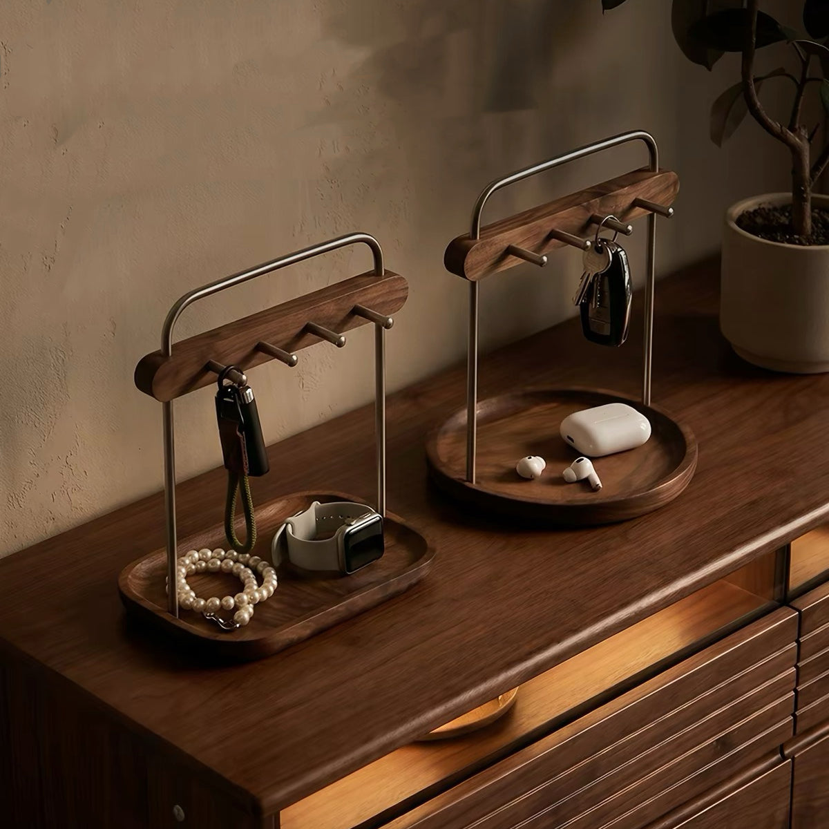 Two wooden key and accessory organizers with metal stands sit on a wooden dresser in a warm, styled room. The left organizer holds keys, a beaded bracelet, and a smartwatch, while the right organizer holds keys and earbuds in their charging case.