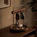 A wooden and metal key holder tray sits on a wooden table in a warm, dimly lit room. The holder has a round wooden base, a vertical metal stand, a top handle, and a horizontal bar holding two keys and a car key fob.