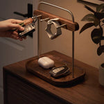 A wooden and metal key holder and organizer tray sits on a wooden table in a warm indoor setting. A hand is holding a key fob near the top rack, and other keys, a white earbud case, and a smartwatch strap are arranged on the holder.