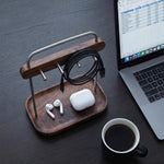 A wooden and metal cable organizer tray sits on a dark desk, holding a black charging cable, white earbuds, and a white charging case. A laptop is partially visible on the right, with a black coffee cup in the foreground.