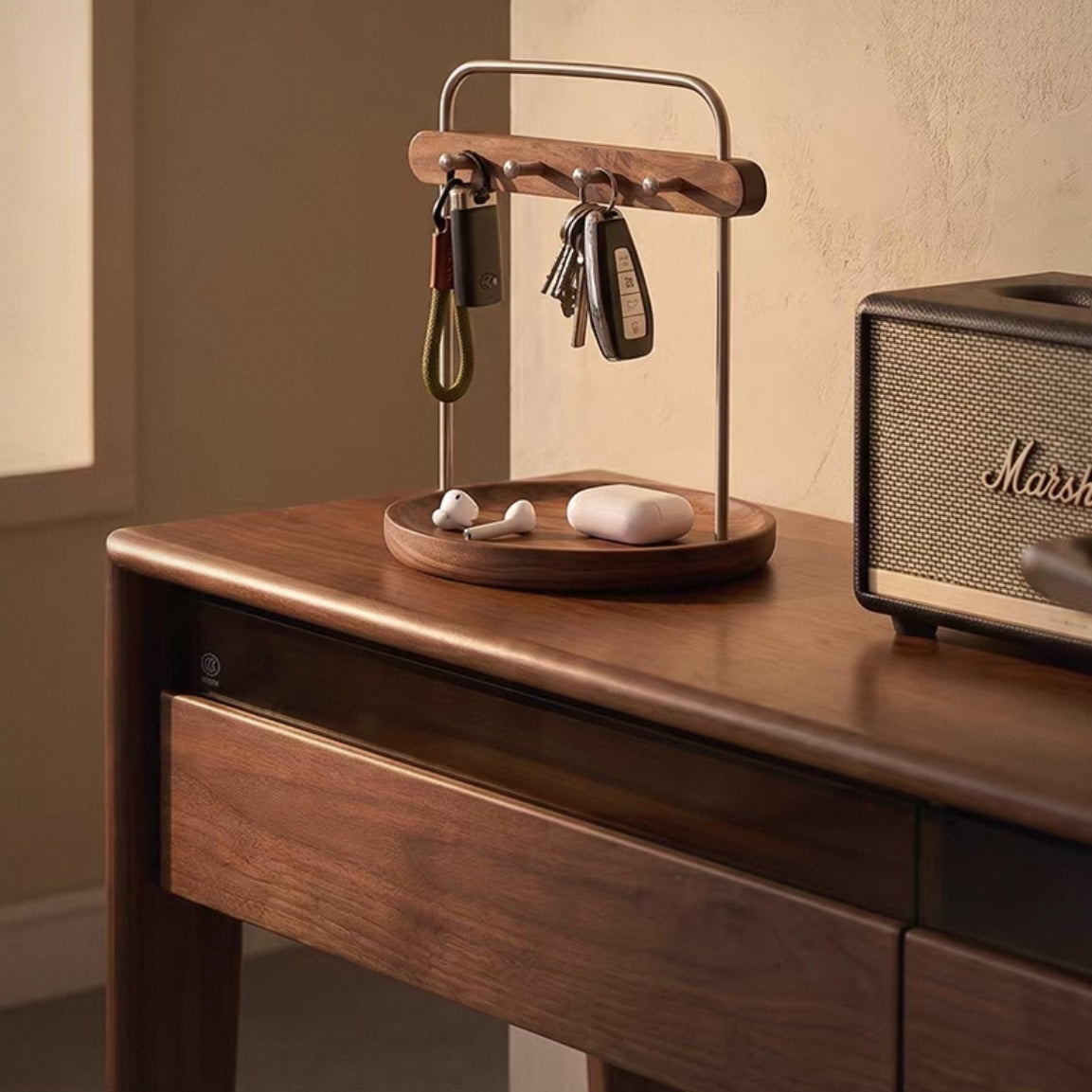 A wooden and metal key holder with hanging keys and a small tray holding earbuds, styled on a wooden desk next to a Marshall speaker.