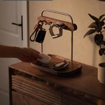 A wooden and metal key holder and tray stand on a wooden cabinet in a warm indoor setting. A hand is placing a white wireless earbud into the tray, with keys and a smartwatch hanging from the metal rack above.