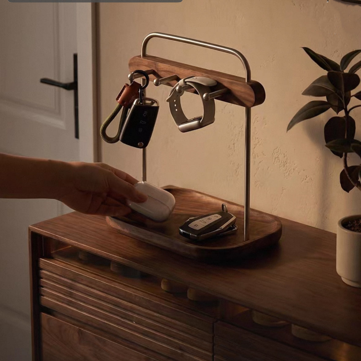 A wooden and metal key holder and tray stand on a wooden cabinet in a warm indoor setting. A hand is placing a white wireless earbud into the tray, with keys and a smartwatch hanging from the metal rack above.
