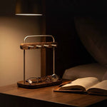 A wooden and metal bedside organizer tray holding eyeglasses and a watch, illuminated by a warm lamp on a wooden table next to an open book in a dimly lit bedroom.