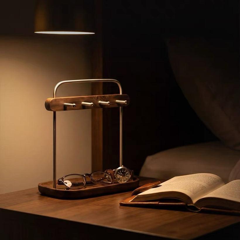 A wooden and metal bedside organizer tray holding eyeglasses and a watch, illuminated by a warm lamp on a wooden table next to an open book in a dimly lit bedroom.