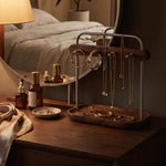 A wooden jewelry organizer with a metal frame stands on a dark wood vanity table in a bedroom setting. The organizer holds several necklaces, rings, and earrings, with a round mirror behind it reflecting a bed and bedside bottles.