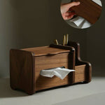 A dark brown wooden tissue box holder with a drawer-style front and open side compartments, shown on a tabletop against a neutral wall. A hand is visible in a circular inset holding part of the product, which also reveals white tissue paper tucked into the holder.