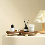 A styled interior scene showing a wooden tray with rounded feet holding a small perfume bottle, a reed diffuser, a candle, and a small box, placed on a pedestal beside a pleated table lamp against a warm beige wall.