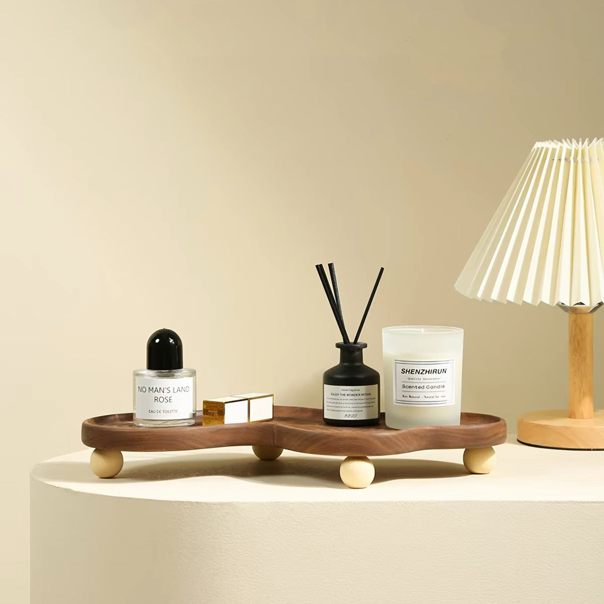 A styled interior scene showing a wooden tray with rounded feet holding a small perfume bottle, a reed diffuser, a candle, and a small box, placed on a pedestal beside a pleated table lamp against a warm beige wall.