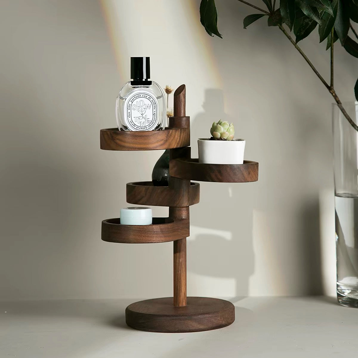 A dark brown wooden spiral shelf tower with multiple tiers, styled with a perfume bottle, a small succulent, and a candle, set against a warm beige wall with a soft spotlight.