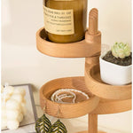 A light natural wood three-tier jewelry organizer with round trays, styled with a candle, a small succulent, and earrings, set against a warm cream background.