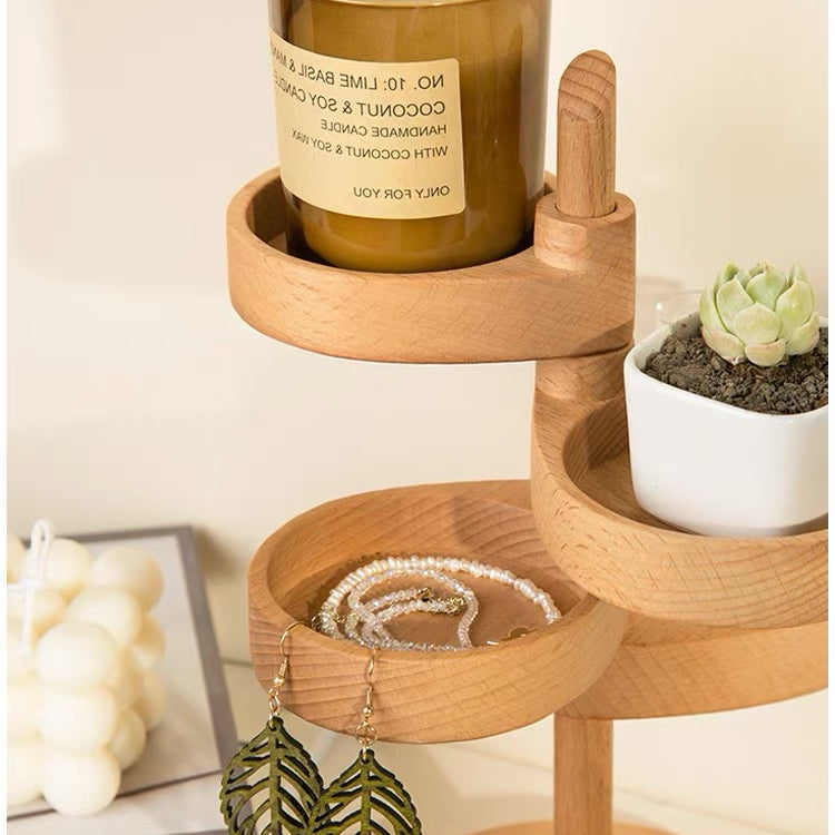 A light natural wood three-tier jewelry organizer with round trays, styled with a candle, a small succulent, and earrings, set against a warm cream background.