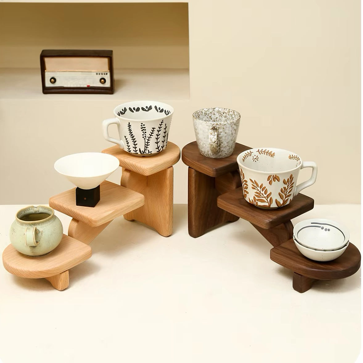 A set of wooden cup and saucer stands in two tones of wood, styled with ceramic mugs and a small lidded container on a warm beige background.