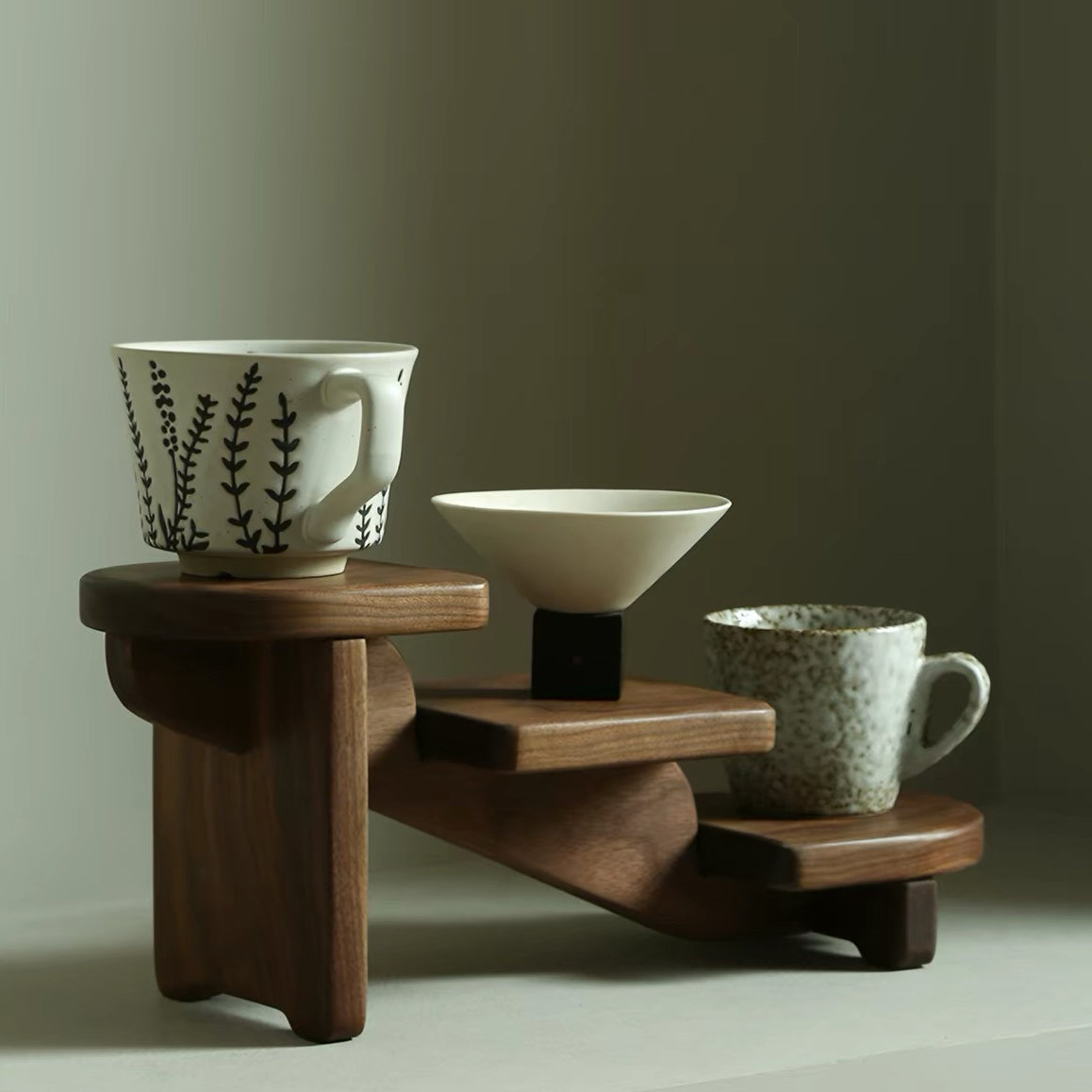 A three-tier wooden coffee station with a dark brown finish, styled with a white ceramic mug featuring black botanical patterns, a cream-colored conical bowl on a small black pedestal, and a speckled gray ceramic mug, set against a neutral warm gray background.