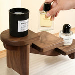 A dark wood multi-tiered shelf organizer holding a black candle, a hand holding a perfume bottle, and a clear bottle with a black cap. The background is a clean beige wall and light wood floor.