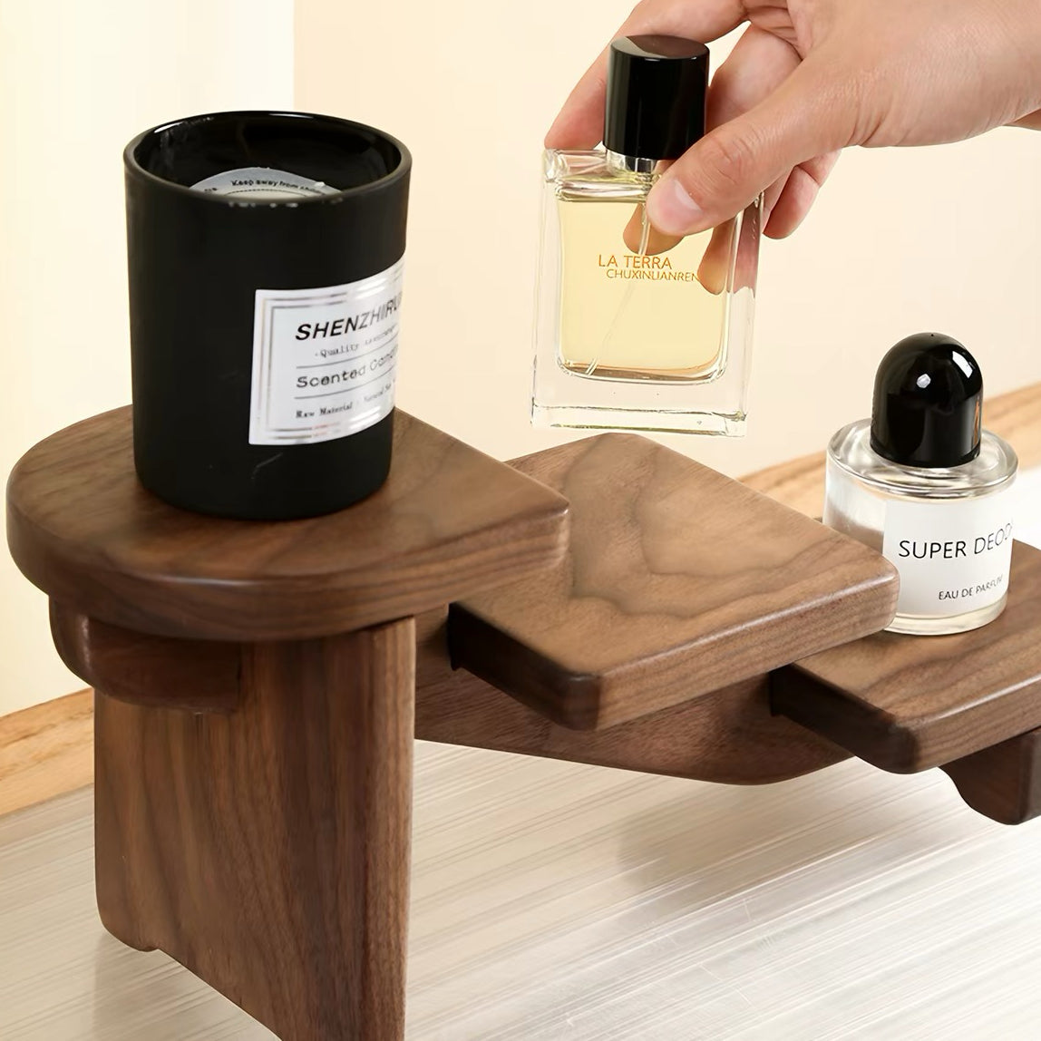 A dark wood multi-tiered shelf organizer holding a black candle, a hand holding a perfume bottle, and a clear bottle with a black cap. The background is a clean beige wall and light wood floor.