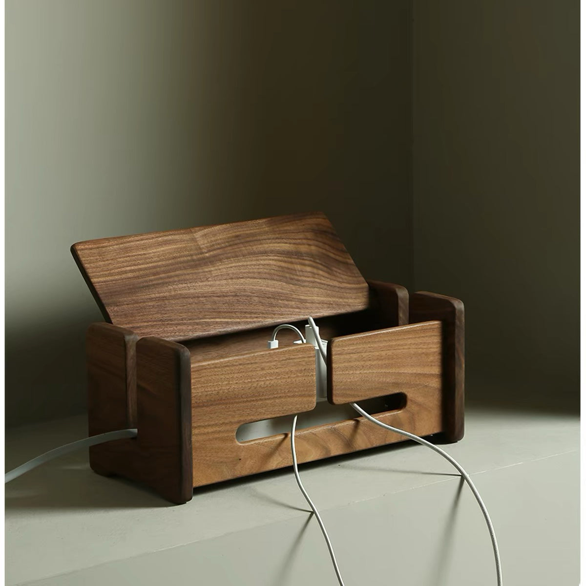 A wooden cable management box with a hinged lid, shown open with white charging cables inserted. The box is made of dark brown wood with a visible grain pattern and has cutouts for cables.