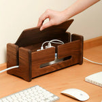 A dark brown wooden desk organizer with a lift-top design, shown on a wooden desk with a hand lifting the lid to reveal storage compartments filled with cables. A keyboard, mouse, and notebook are visible in the foreground.