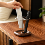 A close-up of a hand placing a white remote control into a silver metal TV stand with a dark wood base, alongside a black remote, set on a wooden table.