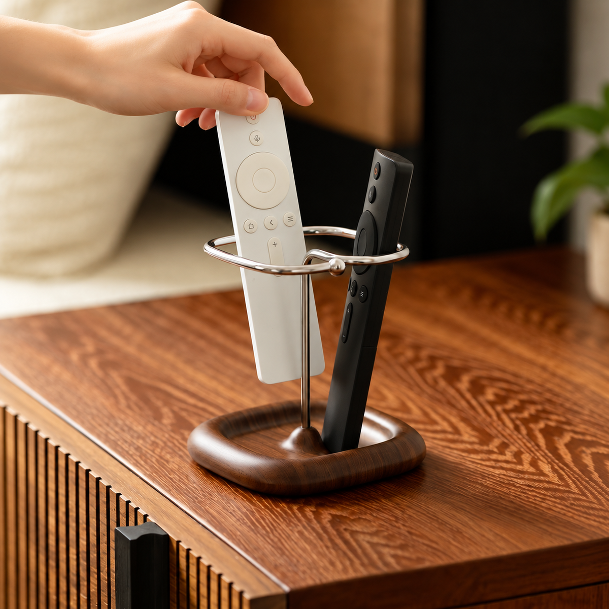 A close-up of a hand placing a white remote control into a silver metal TV stand with a dark wood base, alongside a black remote, set on a wooden table.