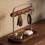 A wooden and metal key holder tray with hooks, shown holding two car keys, a leather strap, and a smartwatch charging case on a wooden table against a textured beige wall.