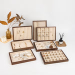 A set of wooden jewelry organizer trays with cream velvet lining, shown open and closed on a white background with decorative props including a vase with dried leaves, pearl beads, and a reed diffuser.