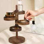 A dark brown wooden multi-tier jewelry organizer stand with a hand placing a ring into one of its bowl compartments. The stand is styled with various cosmetic and jewelry items, set against a warm beige background with a mirror in the background.