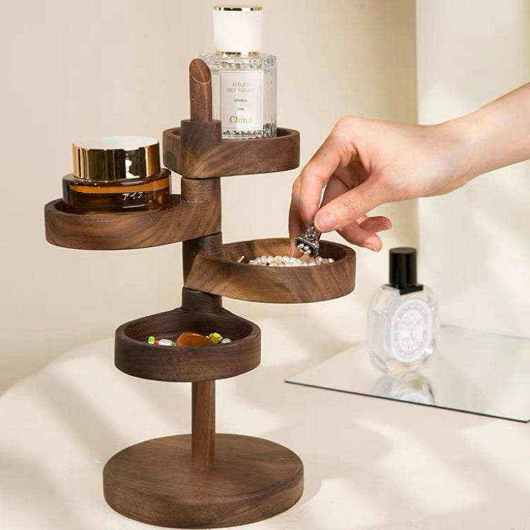 A dark brown wooden multi-tier jewelry organizer stand with a hand placing a ring into one of its bowl compartments. The stand is styled with various cosmetic and jewelry items, set against a warm beige background with a mirror in the background.