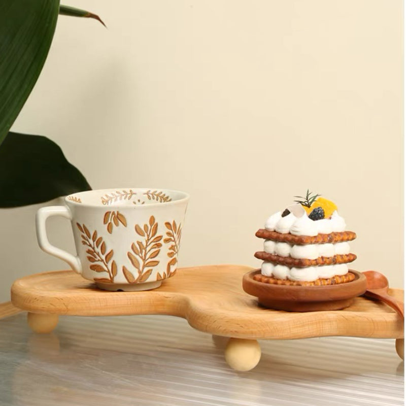 A ceramic mug with a brown leaf pattern on a white base sits on a wooden elevated tray next to a small dessert on a wooden plate, set against a warm beige background with a blurred green leaf in the corner.