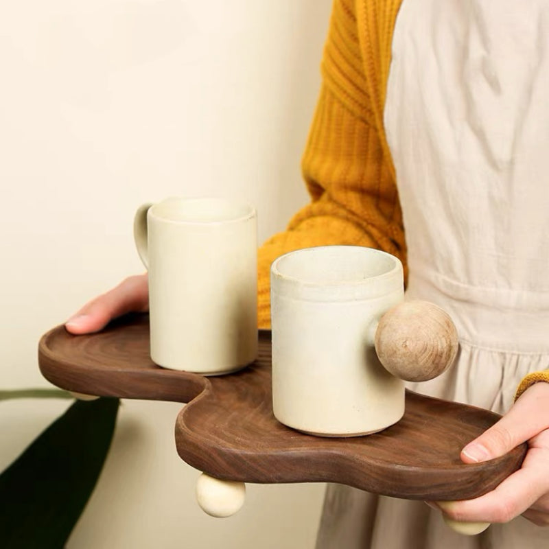 A person in a mustard yellow sweater and white apron holds a dark wood serving tray with two matte ceramic mugs. The tray has organic wavy shapes and small round wooden feet, with one mug featuring a protruding wooden knob.