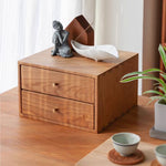 A two-drawer wooden organizer box with a warm medium-brown finish and small round knobs, styled on a wooden table with a white ceramic dish, a gray Buddha figurine, and a plant nearby.