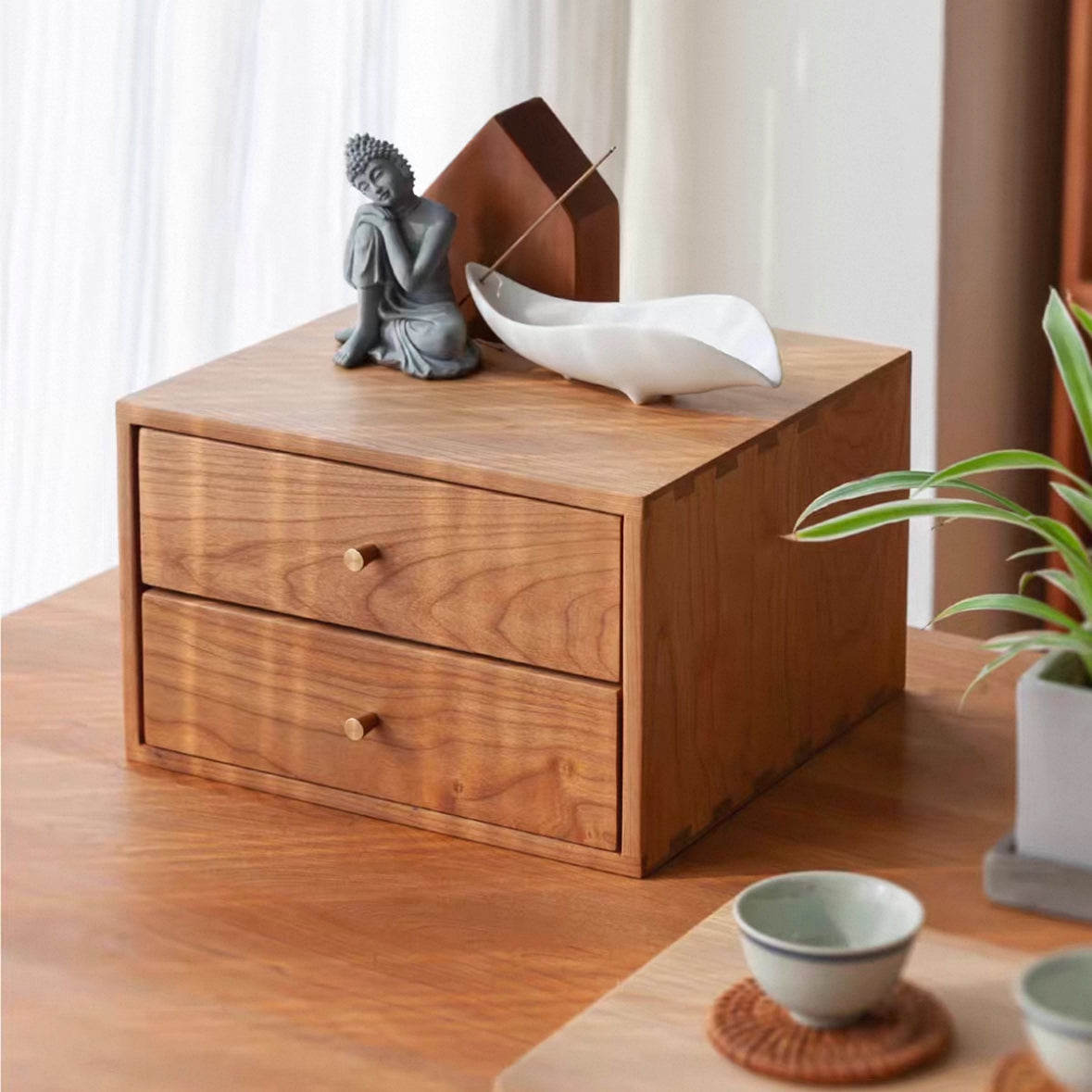 A two-drawer wooden organizer box with a warm medium-brown finish and small round knobs, styled on a wooden table with a white ceramic dish, a gray Buddha figurine, and a plant nearby.