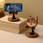 A wooden phone stand shaped like a Shiba Inu dog head, shown on a beige tabletop next to stacked books. One stand is holding a smartphone displaying a dog video call screen, while the other stands empty.