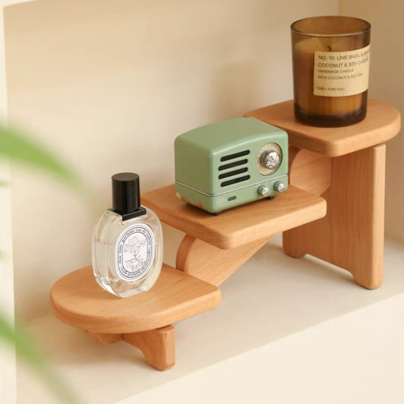A light natural wood step stool or shelf riser holding a mint green retro radio, a clear Diptyque perfume bottle, and an amber glass candle jar, set against a warm cream background.
