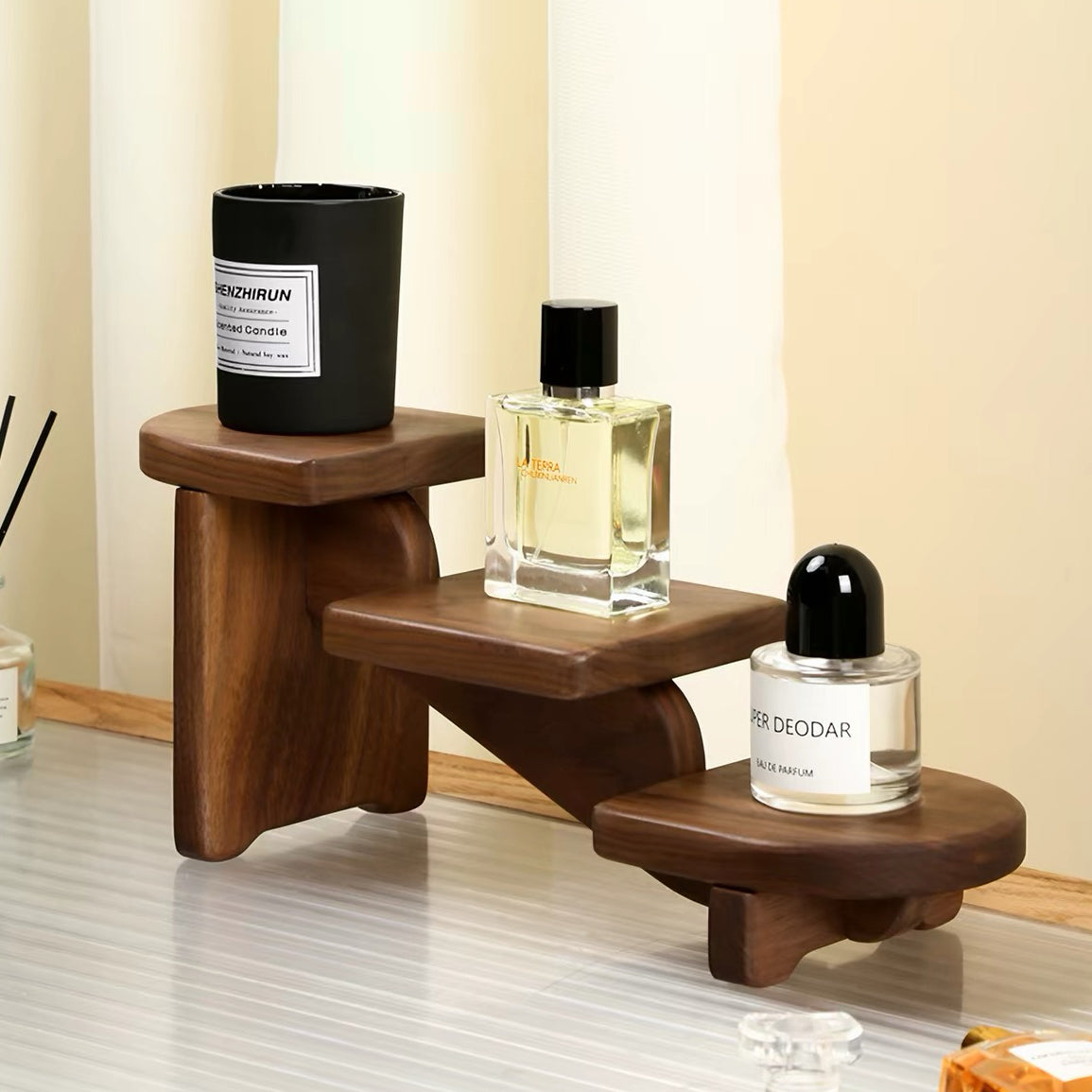 A three-tier wooden step desk organizer holding a black candle jar, a perfume bottle, and a deodorant bottle, set against a warm beige background.