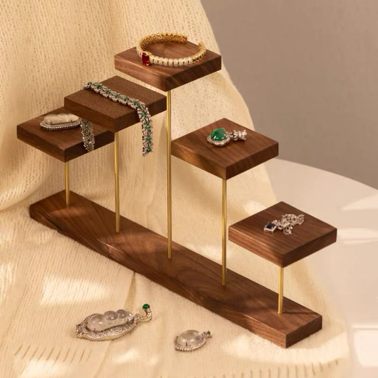 A wooden jewelry display stand with gold-tone metal legs, arranged on a cream textured fabric. The stand has five tiers holding various pieces of jewelry, including bracelets, necklaces, rings, and earrings.