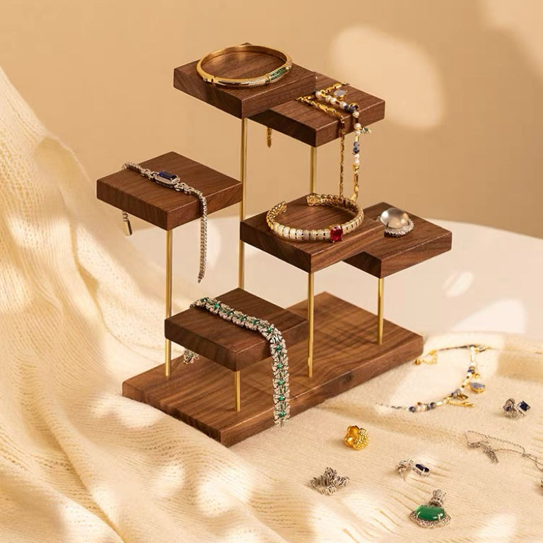 A wooden jewelry display stand with gold-tone metal legs, styled with various gold and silver necklaces, bracelets, and rings on a warm beige fabric background.